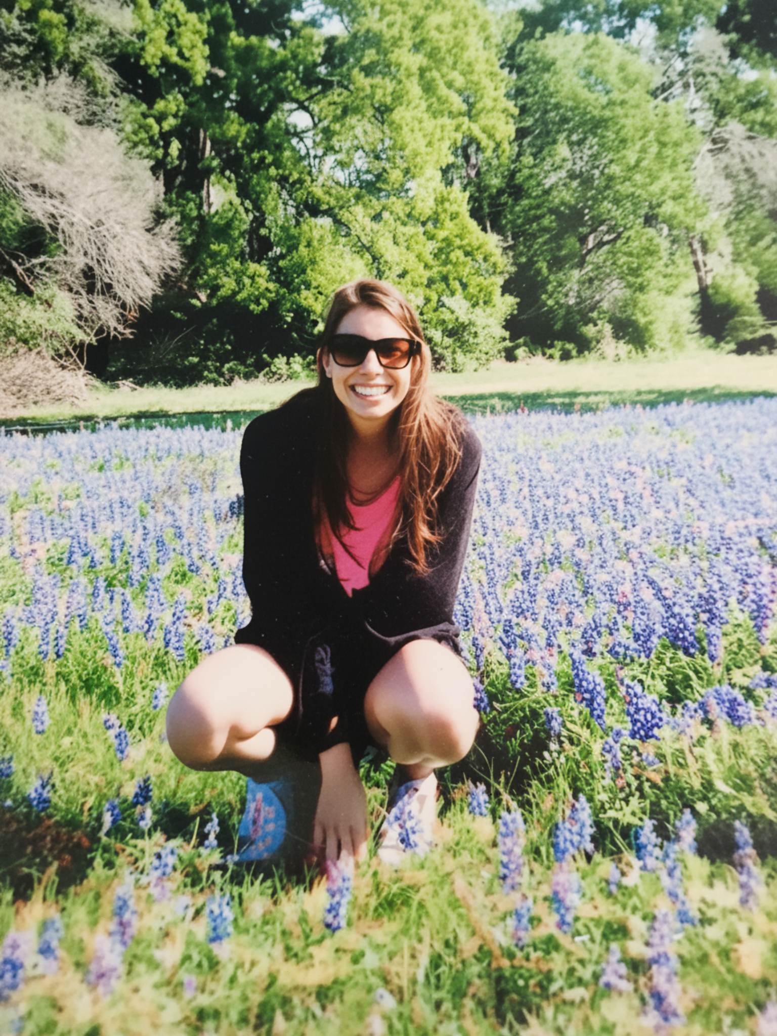 A smiling woman in sunglasses kneeling in a field of Texas bluebonnets